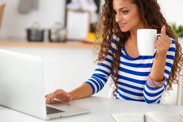 Senyum wanita muda dengan cangkir kopi dan laptop di dapur di rumah — Stok Foto