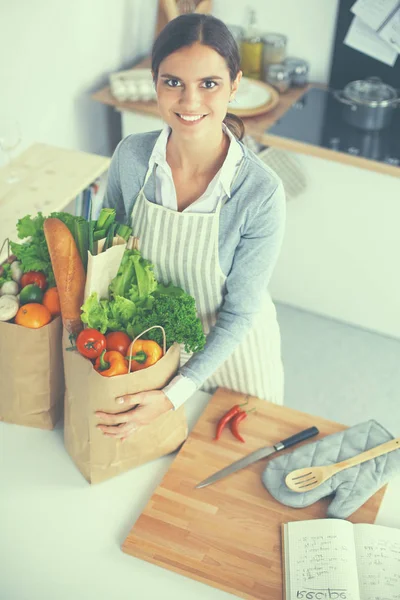 Mujer joven sosteniendo bolsa de la compra de comestibles con verduras. De pie en la cocina —  Fotos de Stock