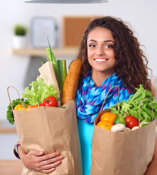 Ung kvinde holder købmand indkøbspose med grøntsager. Stående i køkkenet - Stock-foto