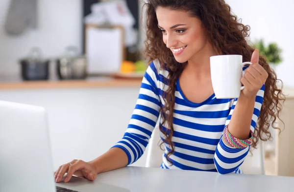 Senyum wanita muda dengan cangkir kopi dan laptop di dapur di rumah — Stok Foto