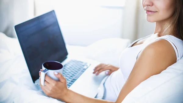 Mujer joven y bonita con portátil en la cama y disfrutando de una taza de té — Foto de Stock