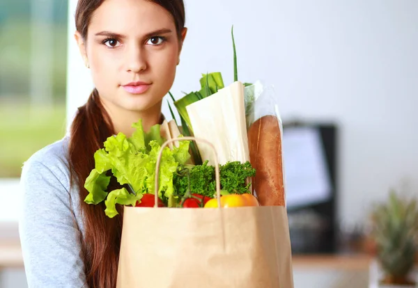 Mujer joven sosteniendo bolsa de la compra de comestibles con verduras. De pie en la cocina — Foto de Stock