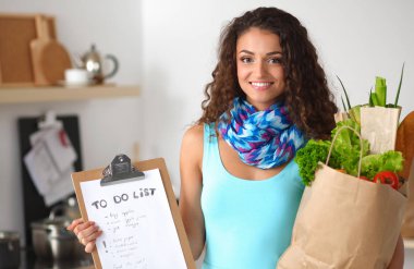 Sebzeli alışveriş poşeti taşıyan genç bir kadın mutfakta duruyor..