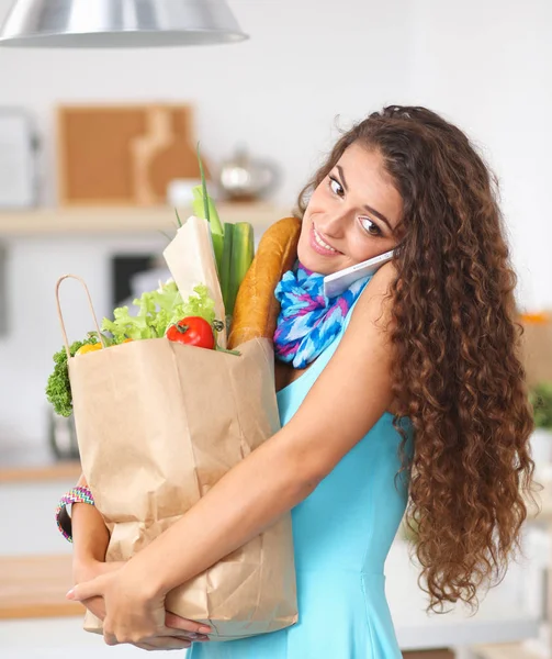 Mujer joven sosteniendo bolsa de la compra de comestibles con verduras de pie en la cocina. — Foto de Stock