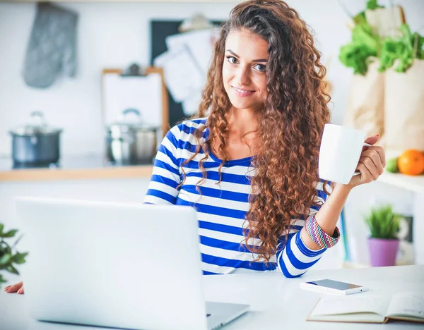 Lachende jonge vrouw met koffiekopje en laptop in de keuken thuis — Stockfoto