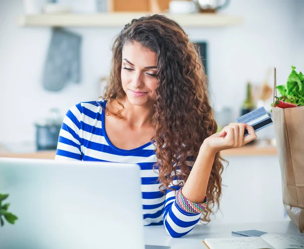 Mujer sonriente compras en línea utilizando la computadora y la tarjeta de crédito en la cocina —  Fotos de Stock