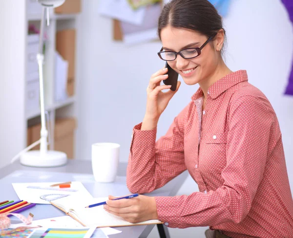 Young attractive female fashion designer working at office desk, drawing while talking on mobile — Stock Photo, Image