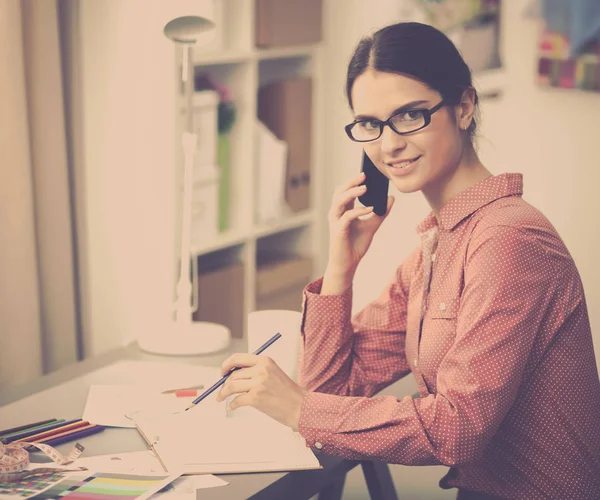 Jonge aantrekkelijke vrouwelijke mode-ontwerper werken op kantoor bureau, tekenen terwijl het praten op mobiele — Stockfoto