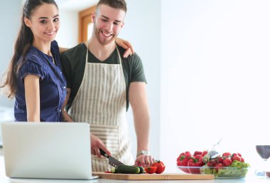 Sebze doğrayan genç bir adam ve mutfakta dizüstü bilgisayarı olan bir kadın.
