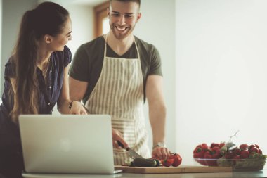 Sebze doğrayan genç bir adam ve mutfakta dizüstü bilgisayarı olan bir kadın.