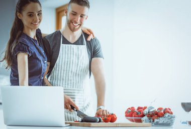 Sebze doğrayan genç bir adam ve mutfakta dizüstü bilgisayarı olan bir kadın.