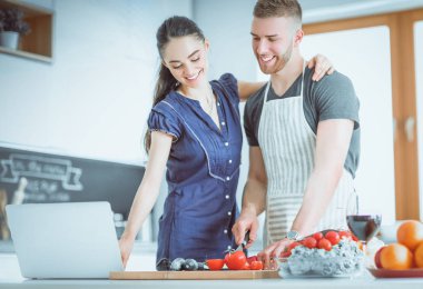 Sebze doğrayan genç bir adam ve mutfakta dizüstü bilgisayarı olan bir kadın.