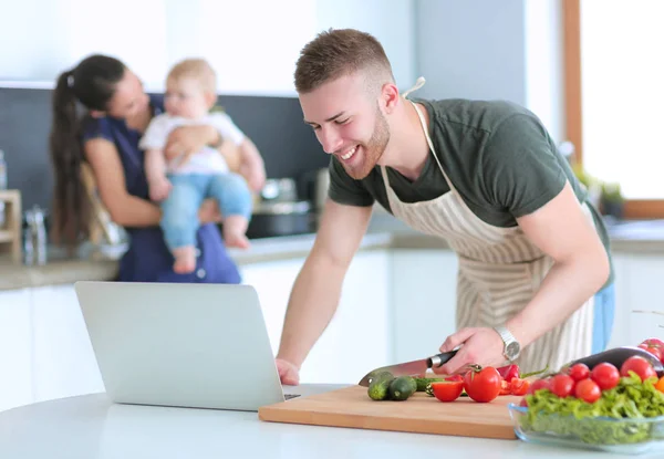 Retrato del hombre cocinando verduras en la cocina mientras mira una computadora portátil en la mesa — Foto de Stock