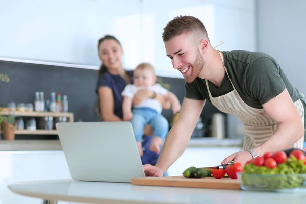 Retrato del hombre cocinando verduras en la cocina mientras mira una computadora portátil en la mesa — Foto de Stock