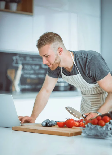 Retrato de homem cozinhando vegetais na cozinha enquanto olha para um computador portátil na mesa — Fotografia de Stock