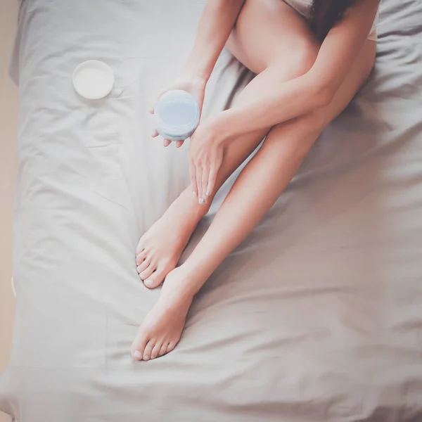 Beautiful woman sitting on bed and applying cream on legs — Stock Photo, Image