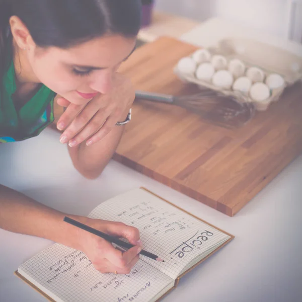 Joyeux belle femme debout dans sa cuisine écrit sur un cahier à la maison — Photo