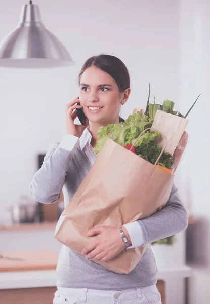 Mujer joven sosteniendo bolsa de la compra de comestibles con verduras. De pie en la cocina — Foto de Stock