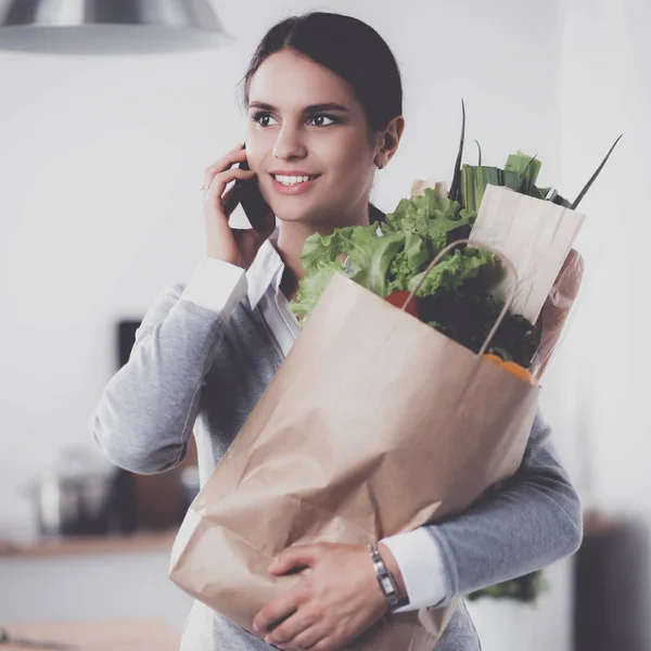 Mujer joven sosteniendo bolsa de la compra de comestibles con verduras. De pie en la cocina —  Fotos de Stock