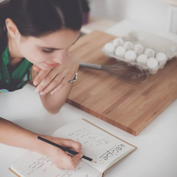 Wanita cantik yang bahagia berdiri di dapur menulis di sebuah buku catatan di rumah — Stok Foto
