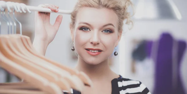 Beautiful young stylist near rack with hangers in office