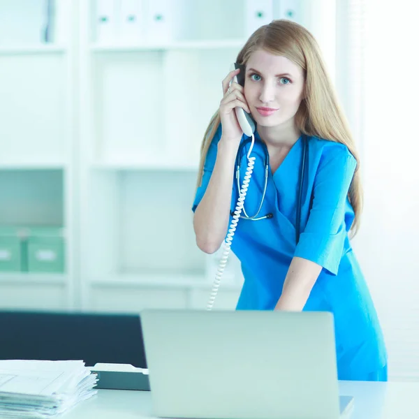 Jonge vrouw arts in witte jas aan de computer met behulp van telefoon — Stockfoto