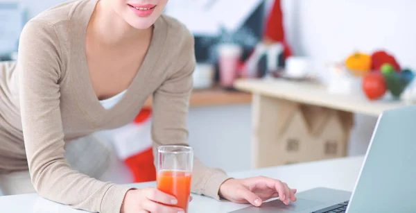 女人在她的厨房里喝果汁的同时使用一台笔记本电脑 — 图库照片