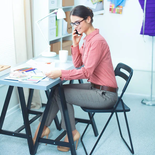 तरुण आकर्षक महिला फॅशन डिझायनर ऑफिस डेस्कवर काम करीत आहे, मोबाइलवर बोलत असताना रेखाचित्र — स्टॉक फोटो, इमेज