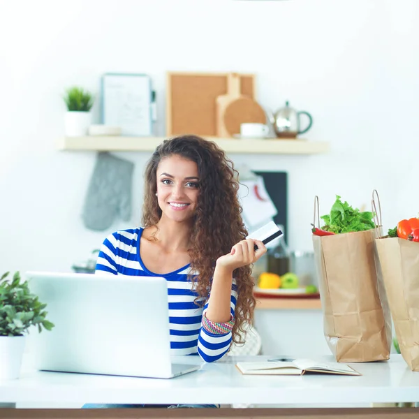 Mujer joven sonriente con taza de café y portátil en la cocina en casa —  Fotos de Stock