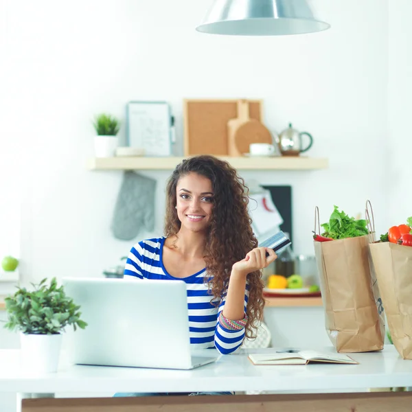 Mujer joven sonriente con taza de café y portátil en la cocina en casa —  Fotos de Stock