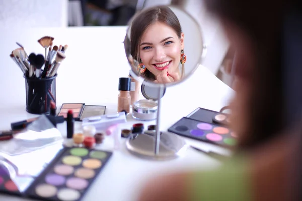 Joven hermosa mujer haciendo maquillaje cerca del espejo, sentado en el escritorio —  Fotos de Stock