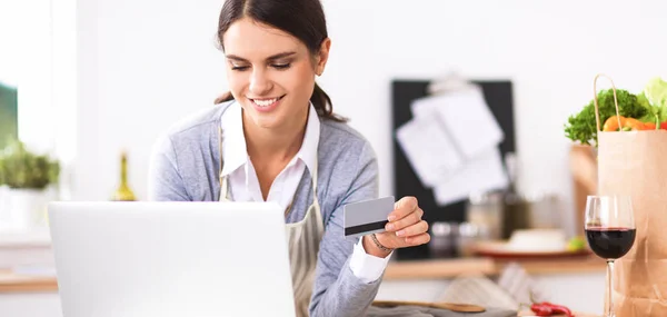 Mujer sonriente compras en línea utilizando la computadora y la tarjeta de crédito en la cocina —  Fotos de Stock