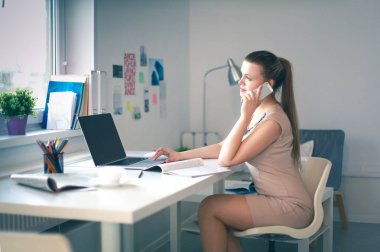 Genç iş kadını masada oturuyor ve telefonla konuşuyor.