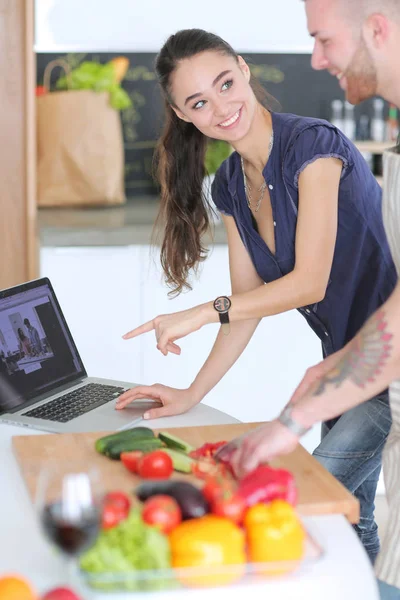 Hombre joven cortando verduras y mujer de pie con el ordenador portátil en la cocina — Foto de Stock