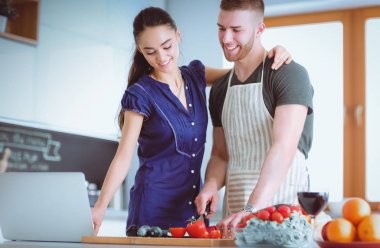 Sebze doğrayan genç bir adam ve mutfakta dizüstü bilgisayarı olan bir kadın.