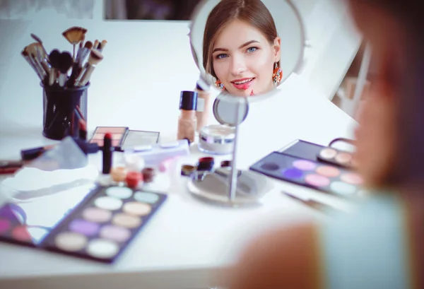 Joven hermosa mujer haciendo maquillaje cerca del espejo, sentado en el escritorio —  Fotos de Stock