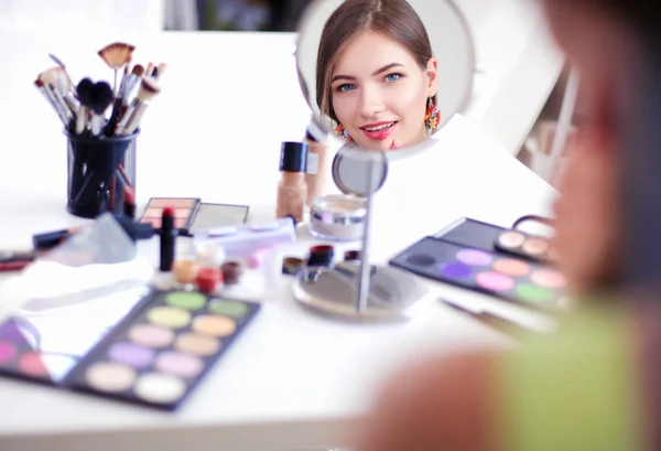 Joven hermosa mujer haciendo maquillaje cerca del espejo, sentado en el escritorio —  Fotos de Stock