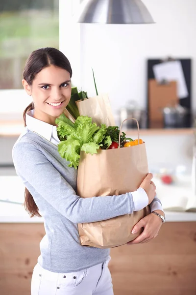 Mujer joven sosteniendo bolsa de la compra de comestibles con verduras. De pie en la cocina — Foto de Stock