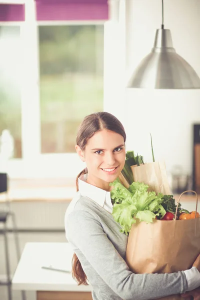 Mujer joven sosteniendo bolsa de la compra de comestibles con verduras. De pie en la cocina —  Fotos de Stock