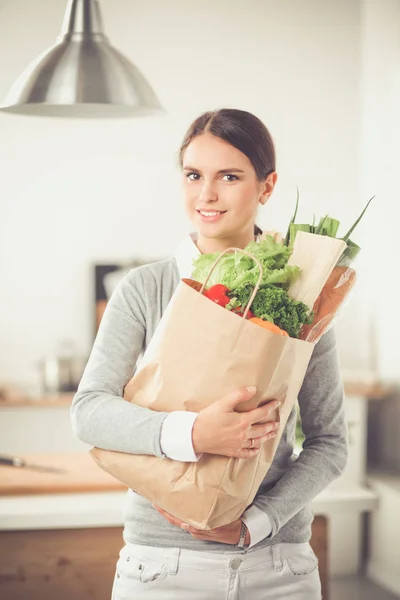 Wanita muda memegang tas belanja dengan sayuran. Berdiri di dapur — Stok Foto