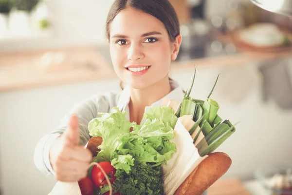 Młoda kobieta trzyma torbę na zakupy z warzywami. Stojąc w kuchni — Zdjęcie stockowe