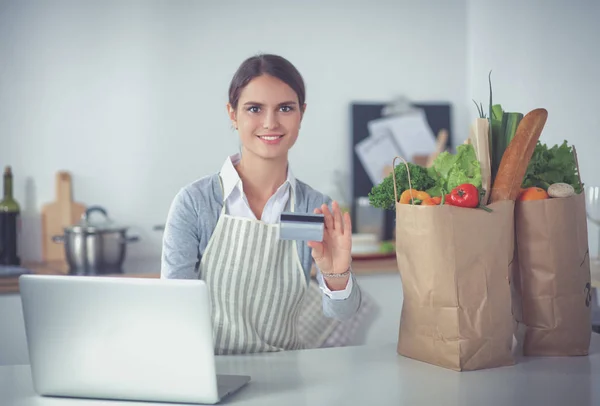 Mujer sonriente compras en línea utilizando la computadora y la tarjeta de crédito en la cocina — Foto de Stock