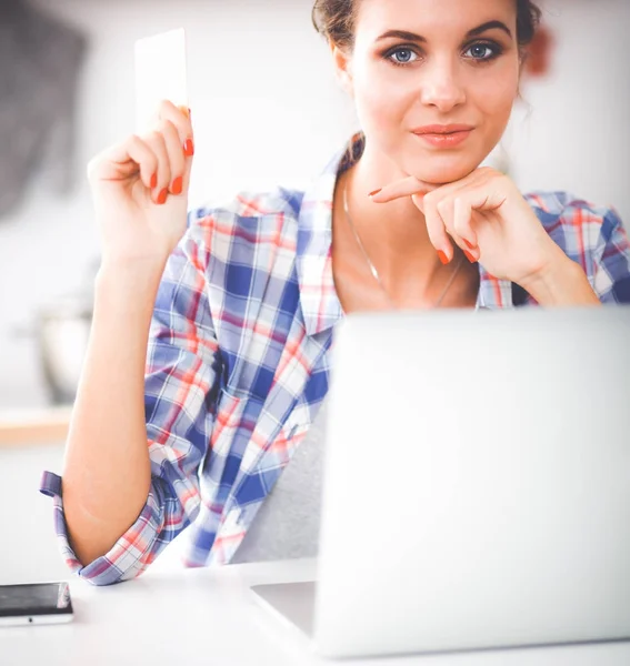 Mujer sonriente compras en línea utilizando la computadora y la tarjeta de crédito en la cocina —  Fotos de Stock