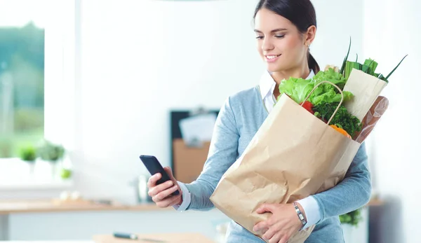 Mujer joven sosteniendo bolsa de la compra de comestibles con verduras. De pie en la cocina —  Fotos de Stock