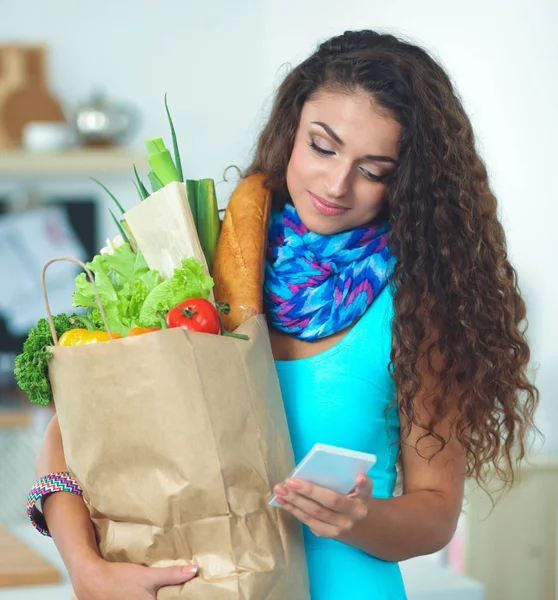 Uśmiechnięta kobieta z telefonem komórkowym trzymająca torbę na zakupy w kuchni — Zdjęcie stockowe