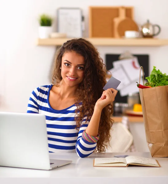 Mujer sonriente compras en línea utilizando la computadora y la tarjeta de crédito en la cocina —  Fotos de Stock