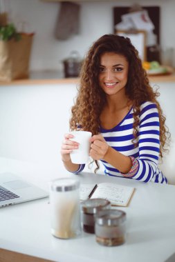 Kahve fincanı ve dizüstü evde mutfakta gülümseyen genç kadınla. Gülümseyen genç kadın