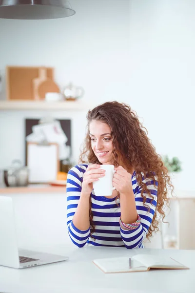 Lachende jonge vrouw met koffiekopje en laptop in de keuken thuis. Lachende jonge vrouw — Stockfoto
