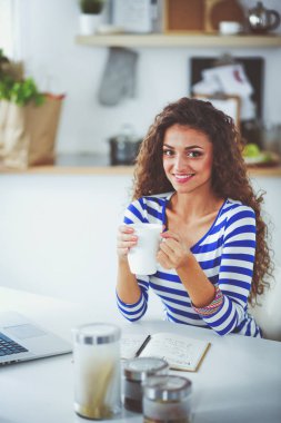Kahve fincanı ve dizüstü evde mutfakta gülümseyen genç kadınla. Gülümseyen genç kadın