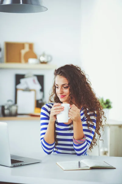 Mujer joven sonriente con taza de café y portátil en la cocina en casa. Jovencita sonriente —  Fotos de Stock